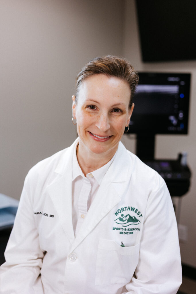 Smiling healthcare professional in a white lab coat in a medical office, with a monitor in the background.
